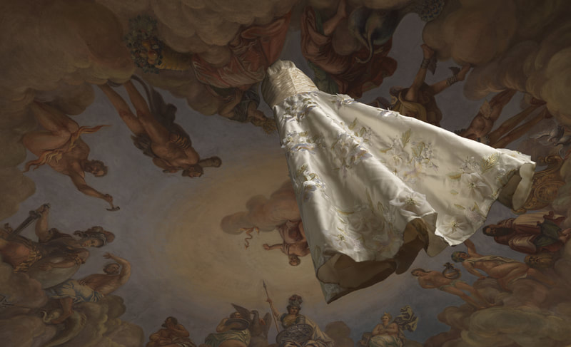 Ein weisses Hochzeitskleid hängt von einer Decke, die mit einem Götterhimmel bemalt ist.