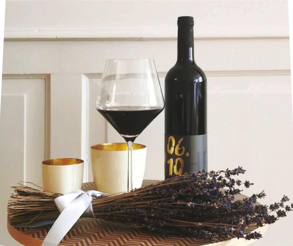 Auf einem kleinen runden Tischchen aus Holz stehen eine Weinflasche, ein Glas Rotwein, zwei Windlichter und ein Bündel Lavendel, das von einer blass lila Schleife zusammengehalten wird.